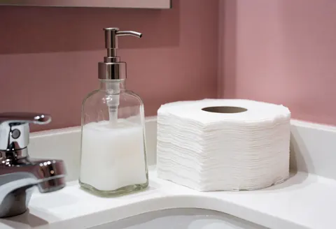 Comptoir de salle de bain équipé d'un distributeur de savon, de serviettes en papier, d'un robinet chromé et d'un mur rose, idéal pour un salon de coiffure ou de beauté.