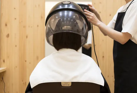 Une cliente sous un grand séchoir avec une serviette blanche tandis qu'un coiffeur ajuste les commandes dans un salon aux murs clairs.