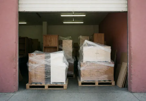 Des meubles en bois enveloppés dans du plastique sont alignés dans un entrepôt aux murs roses avec une porte ouverte.