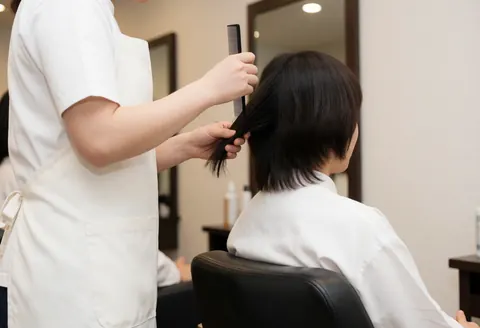 Un coiffeur en tablier blanc utilise une peigne à raton pour séparer les cheveux d'un client assis sur une chaise noire dans un salon lumineux aux murs neutres.