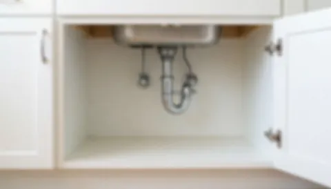 Vue rapprochée d'une armoire blanche sous un évier, avec des tuyaux visibles en arrière-plan flou, servant de fond neutre pour du texte commercial.