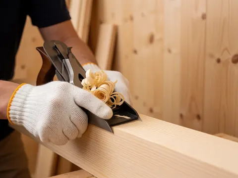 Gros plan montrant une main gantée travaillant le bois de pin avec des copeaux visibles autour du tablier de travail.