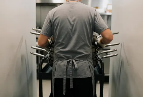 Un cuisinier professionnel charge des casseroles sur un chariot dans une cuisine industrielle avec des murs gris en arrière-plan flou.