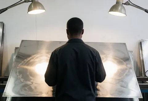Un ouvrier de dos examine une grande plaque d'acier inoxydable dans un atelier industriel éclairé par des lumières de travail sur fond blanc.