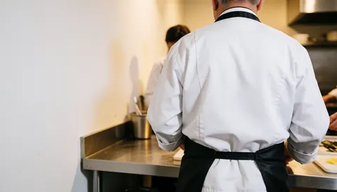 Vue arrière d'un chef cuisinier travaillant dans une cuisine professionnelle avec espace de copie à gauche pour texte, ambiance réaliste et propre.