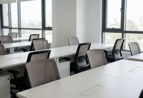 Vue d'un espace de coworking moderne et vide, comprenant un bureau, une chaise ergonomique, une fenêtre laissant entrer la lumière naturelle et une vue sur un bâtiment extérieur.