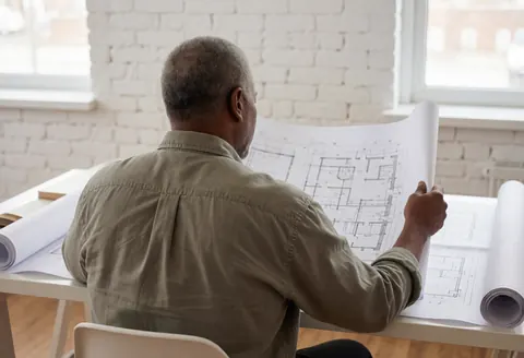 Un architecte consulte des plans d'architecture dans un bureau moderne aux murs de briques blanches, baigné de lumière naturelle et offrant une ambiance professionnelle.