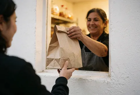 Une propriétaire de boulangerie souriante tend un sac alimentaire à un client par une fenêtre, avec une lumière chaleureuse et un mur blanc en arrière-plan.