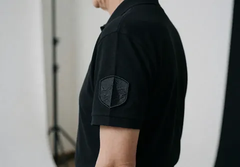 Homme d'affaires en studio sur fond blanc, vêtu d'un polo noir, mettant en valeur le détail du patch brodé et la texture du tissu sous un éclairage soigné.