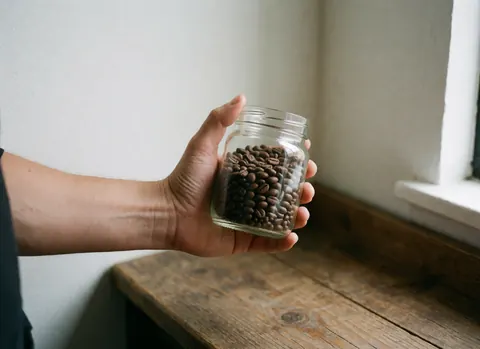 Des grains de café frais sont disposés dans une jarre en verre posée sur une surface en bois, baignés par la lumière naturelle provenant d'une fenêtre.