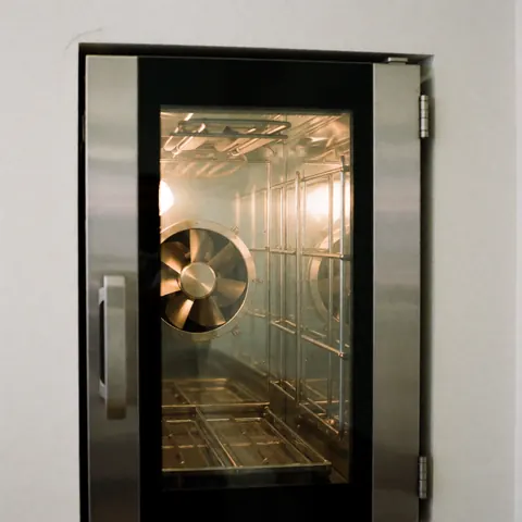 Intérieur d'un four de convection industriel avec ventilateur et éléments chauffants visibles à travers la porte en verre, sur fond blanc.