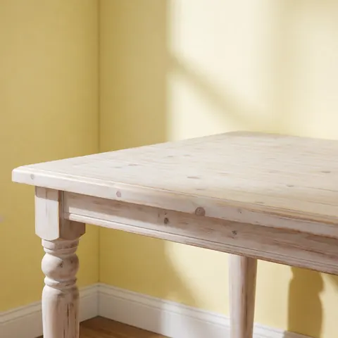 Table en bois de pin au style rustique, peinte en blanc délavé, posée sur une surface claire devant un mur jaune pâle éclairé par une lumière matinale douce.