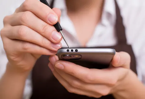Mains d'un technicien en train de réparer un smartphone à l'aide d'outils, sur fond flou, illustrant un service technique en action.