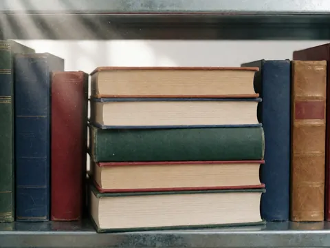 Une pile de livres aux couvertures colorées posée sur une étagère en métal, éclairée par un rayon de lumière mettant en valeur la poussière.