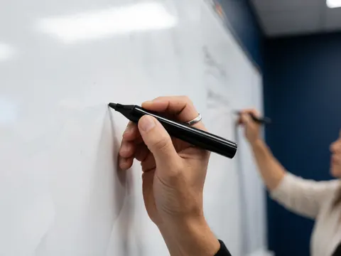 Une main portant une bague écrit avec un marqueur noir sur un tableau blanc, une autre personne en arrière-plan, mur bleu foncé, ambiance de classe ou réunion professionnelle.