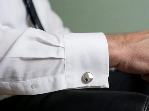 Gros plan d'une manchette de chemise blanche avec des boutons de manchette argentés posés sur du cuir noir, sur un fond vert pâle, évoquant le professionnalisme et la précision.