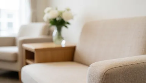 Vue d'un intérieur lumineux et apaisant d'une salle d'attente, comprenant un canapé beige confortable, une table en bois et un vase de fleurs blanches près d'une fenêtre.