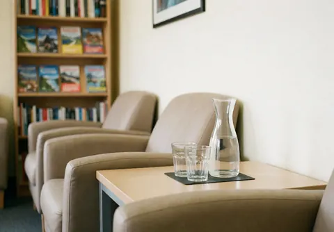 Vue latérale d'une salle d'attente propre et lumineuse équipée de chaises, d'une table basse en bois, de verres et d'étagères remplies de livres.