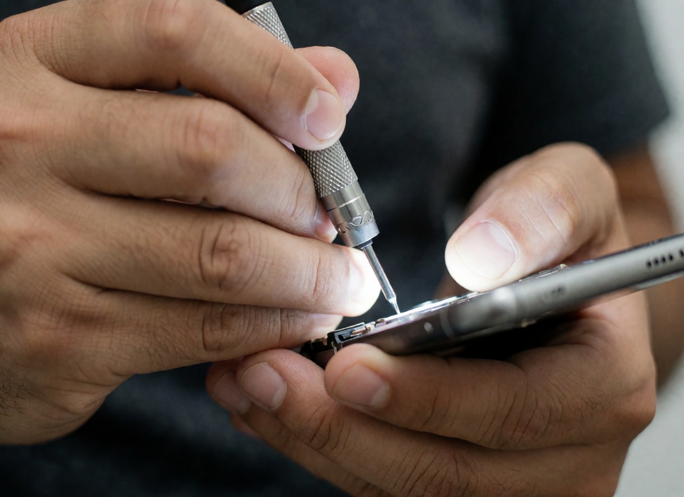 Gros plan sur les mains d'un technicien utilisant un tournevis de précision pour démonter un smartphone, mettant en évidence les détails techniques et l'outil sur fond flou.