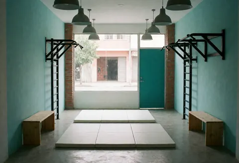 Vue intérieure d'un espace de fitness vide et propre avec murs turquoise, sol en caoutchouc gris, bancs en bois et équipements de sport sous un éclairage naturel.