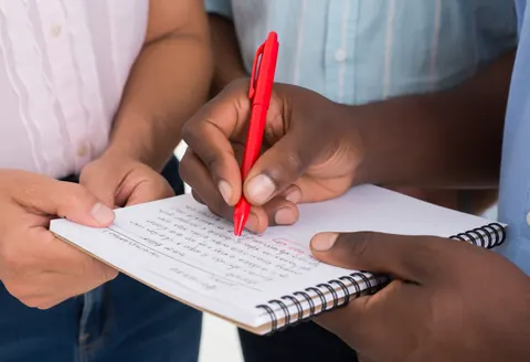 Un professeur corrige un exercice dans un cahier avec un stylo rouge pendant qu'un étudiant observe attentivement.