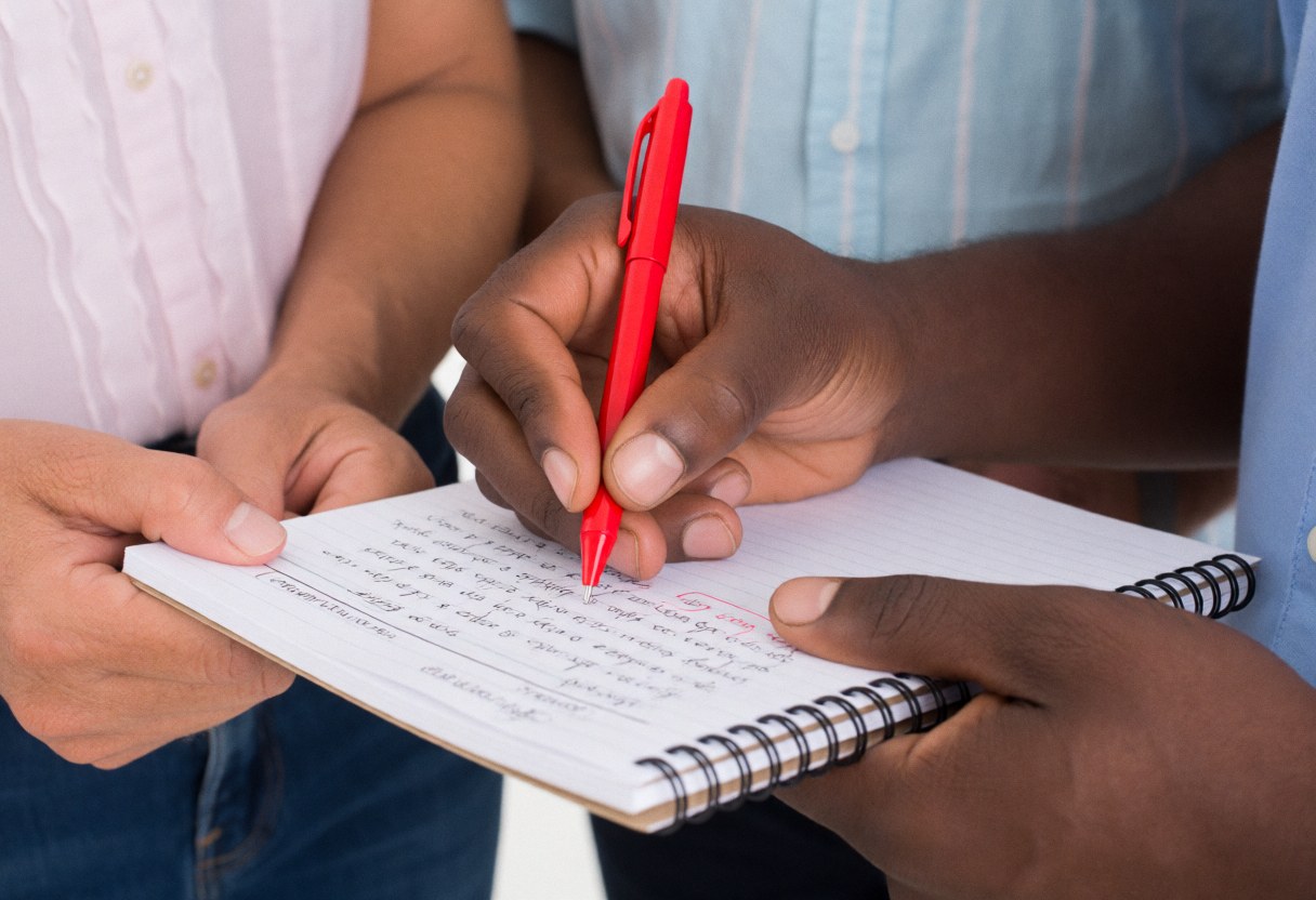 Un professeur corrige un exercice dans un cahier avec un stylo rouge pendant qu'un étudiant observe attentivement.