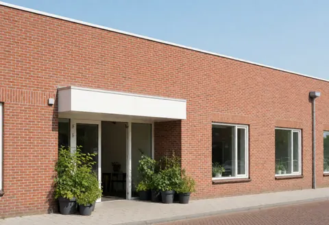 Façade d'un bâtiment communautaire rénové avec mur en brique, entrée sous auvent blanc, plantes en pots, ciel clair et ambiance moderne.