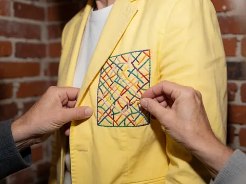 Mains effectuant une broderie colorée sur une veste jaune posée sur un fond de briques, détail artisanal pour le branding d'entreprise.