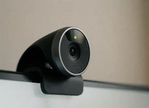 Webcam noir avec voyant vert allumé fixé sur écran, fond beige flou, détail technologique pour réunions en entreprise.