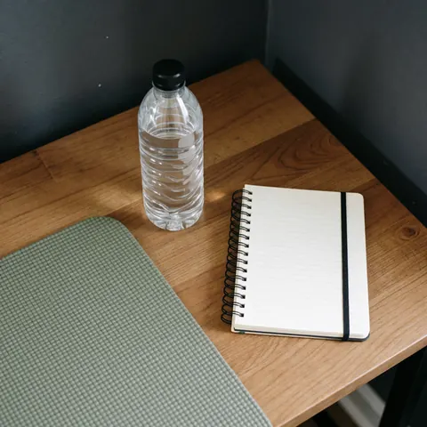 Kit bien-être composé d'une bouteille d'eau, d'un carnet et d'un tapis de yoga disposés sur une table en bois devant un mur gris.