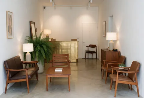 Vue d'un lobby d'hôtel élégant avec mobilier en bois et bronze, murs blancs, plantes vertes et éclairage ambiant créant une atmosphère calme et accueillante.