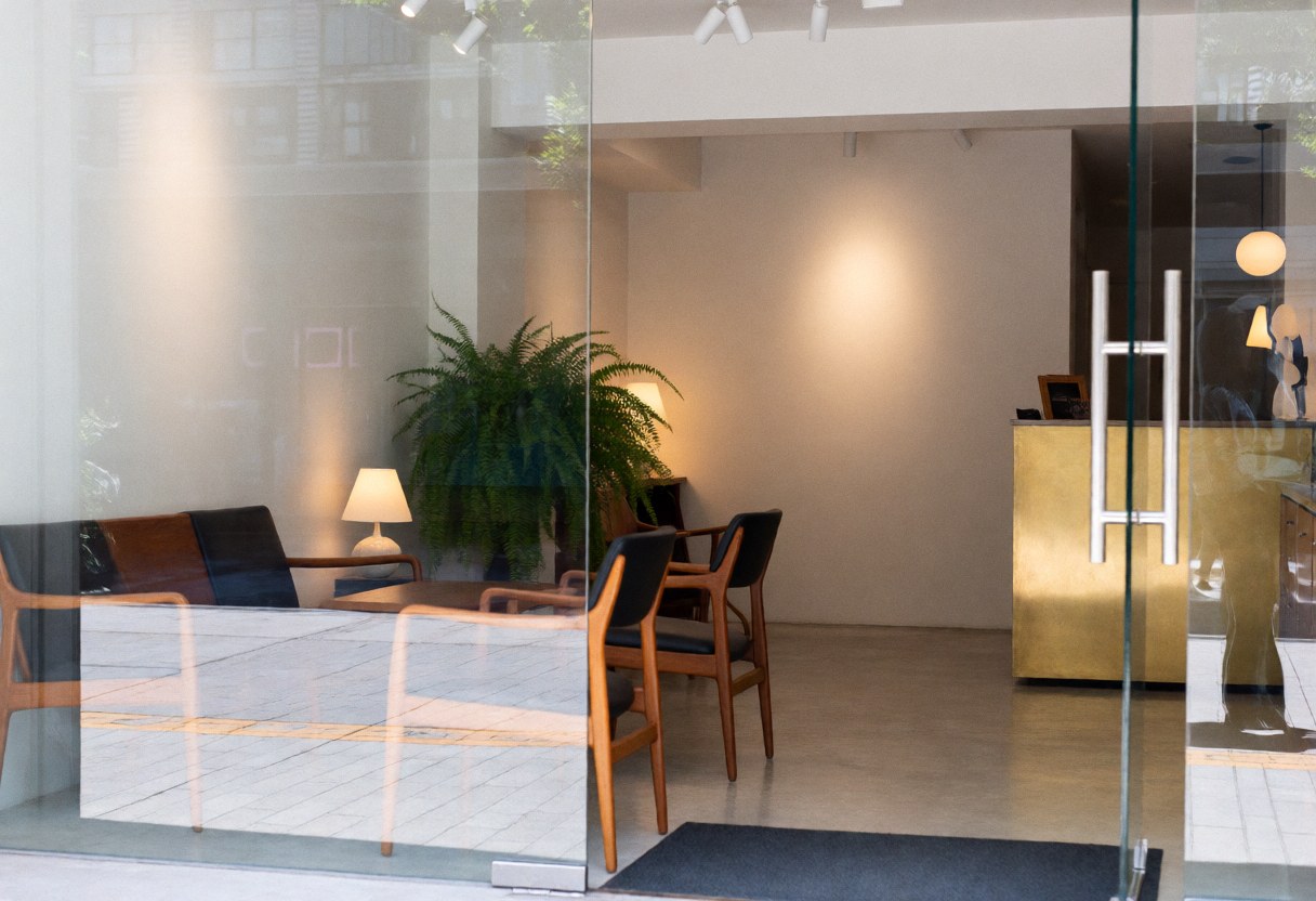 Entrée d'un hôtel boutique avec réception en bronze, meubles en teck, murs blancs et une grande plante à l'intérieur.