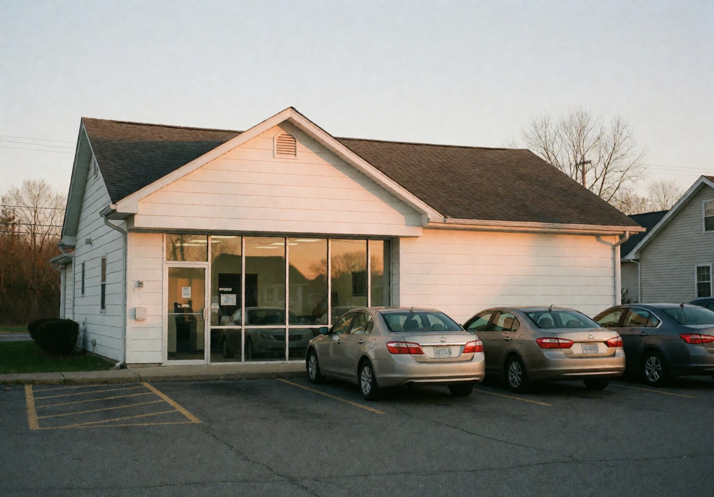 Façade blanche d'une agence d'assurance avec grande fenêtre et voitures stationnées au parking lors du coucher du soleil.