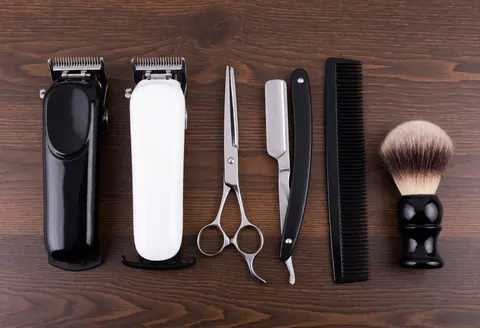Vue de dessus d'outils de barbier incluant ciseaux, rasoir, peigne, brosse et tondeuse sur une surface en bois sombre avec éclairage professionnel.