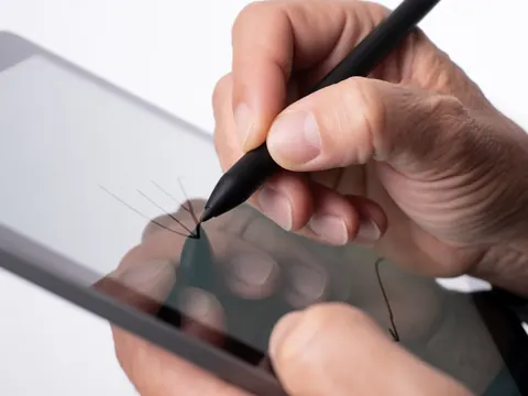 Une main adulte trace des lignes précises avec un stylet noir sur l'écran d'une tablette, révélant un design digital professionnel sous une illumination claire sur fond blanc.