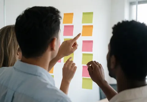 Trois personnes organisent des notes adhésives colorées sur un mur blanc lors d'une réunion professionnelle lumineuse.