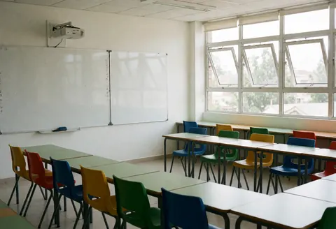 Salle de classe vide équipée d'un tableau blanc, de fenêtres ouvertes laissant entrer la lumière naturelle et de chaises aux couleurs vives. Ambiance lumineuse et moderne.