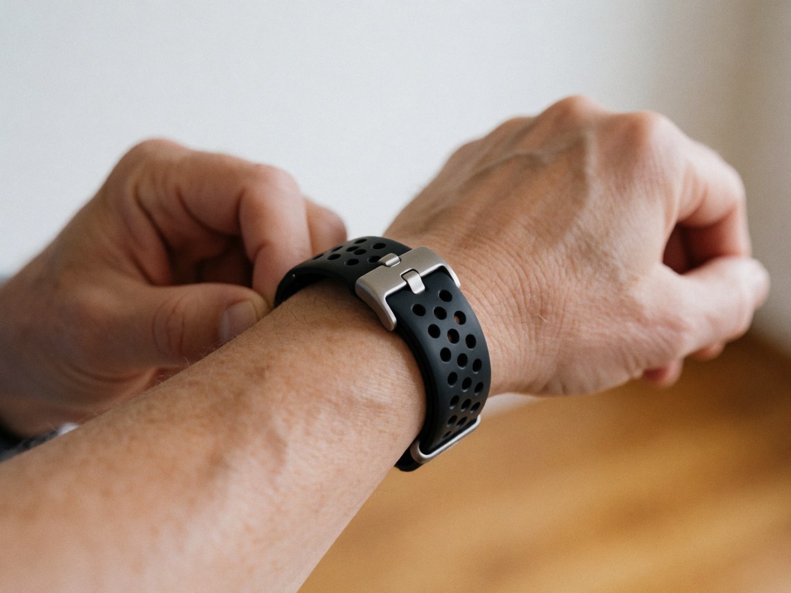 Gros plan sur la boucle d'une montre connectée avec bracelet noir perforé et métal brillant tenue dans une main sur fond bois clair.