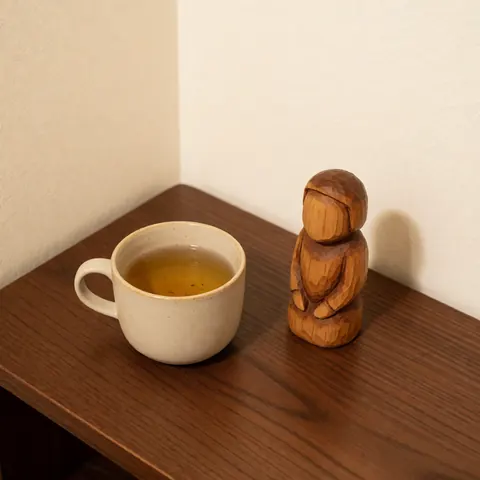 Une tasse de thé et une petite sculpture en bois reposent sur une table sombre dans un intérieur aux murs clairs, évoquant calme et bien-être.