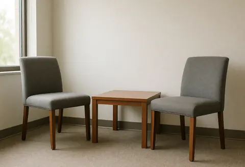 Vue d'une salle de réunion ou d'entretien vide, équipée d'une table en bois et de chaises grises, baignée de lumière naturelle provenant d'une fenêtre.