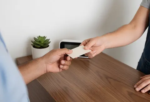 Deux personnes échangent des cartes de visite sur une table en bois avec plantes succulentes et horloge numérique en arrière-plan.