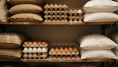 Vue d'un arrière-plan flou montrant des sacs de farine, des œufs et des boîtes sur une étagère en bois dans un intérieur chaleureux.