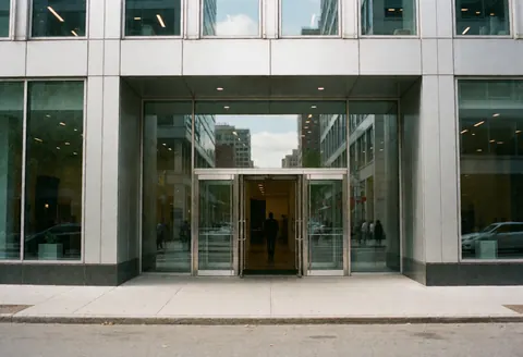 Façade contemporaine en verre reflétant la ville, une personne franchit le seuil principal, évoquant professionnalisme et accès aux entreprises.