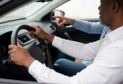 Vue intérieure d'un véhicule montrant un instructeur pointant vers l'avant tandis qu'un élève tient le volant, avec une rue visible par la fenêtre en pleine journée.