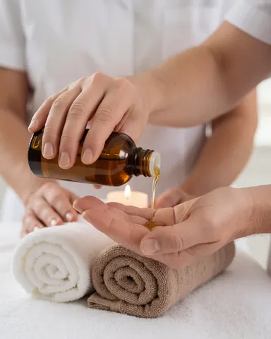 Mains en contact avec de l'huile de massage, serviettes blanches et bougie créant une ambiance spa relaxante.