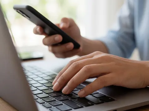 Mains d'une personne travaillant sur un ordinateur portable et tenant un téléphone portable dans un espace de travail professionnel avec éclairage naturel.