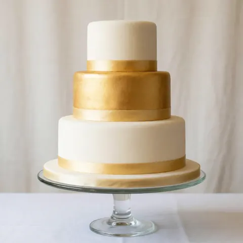 Un gâteau de mariage élégant aux tons pastels orné de détails dorés, posé sur un socle transparent en verre, évoquant une célébration raffinée et festive.