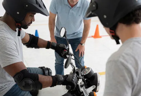Un élève en tenue de protection complète suit une leçon pratique de conduite de moto sous la supervision d'un instructeur dans un parcours délimité par des cônes.