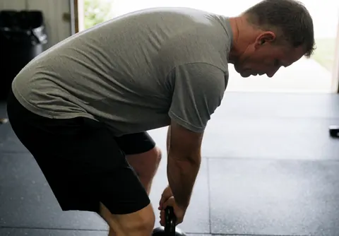 Un homme effectue un exercice de force avec un kettlebell dans une salle de sport équipée d'un sol en caoutchouc et éclairée par la lumière naturelle.