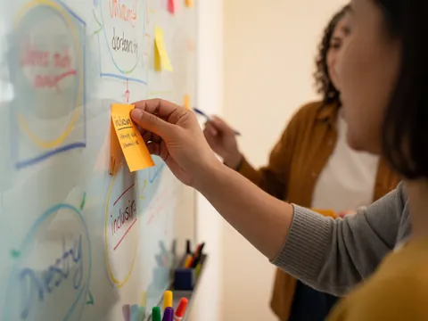 Une main place une note adhésive orange portant le mot inclusion sur un tableau blanc avec des diagrammes, tandis qu'une autre personne écrit en arrière-plan lors d'une session de formation.